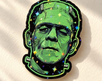 May include: A die-cut sticker featuring a green-skinned monster with a flat head, heavy eyelids, and metal neck bolts. The illustration has a holographic finish with small, colorful star-shaped sparkles scattered across the surface.