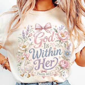 Könnte beinhalten: Ein cremefarbenes Kurzarm-T-Shirt mit einem Grafikdruck einer Ente mit rosa Schleife, umgeben von Blumenillustrationen und einer großen rosa Schleife oben. Der Text lautet God Is Within Her Psalm 46:5 in rosa und blauer Schrift.