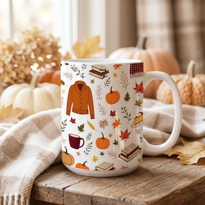 May include: A white ceramic mug featuring a repeating pattern of orange cardigans, pumpkins, stacks of books, slices of pie, and colorful autumn leaves. The design includes various foliage elements in shades of green, yellow, and red, creating a detailed seasonal motif across the exterior surface of the mug.