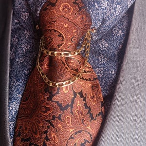 Peut inclure: Gros plan d'une cravate à motifs cachemire marron et noir, maintenue par une pince à cravate dorée. La cravate est portée sur une chemise bleue à motifs et un blazer gris.