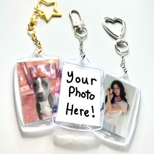 May include: Three clear acrylic photo frame keychains with metal attachments. One features a dog in a hat, one displays the text Your Photo Here!, and one shows a person holding a small animal. The keychains have star, lobster, and heart clasps.