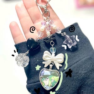 May include: A silver keychain with a heart-shaped clasp, adorned with various charms. Charms include a black heart, a clear flower, a white bow, a star, and a heart-shaped pendant with a character illustration. The keychain features clear and pearl beads.