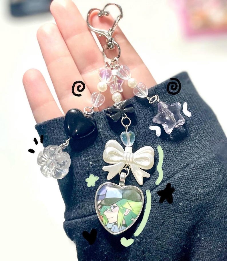 May include: A silver keychain with a heart-shaped clasp, adorned with various charms. Charms include a black heart, a clear flower, a white bow, a star, and a heart-shaped pendant with a character illustration. The keychain features clear and pearl beads.