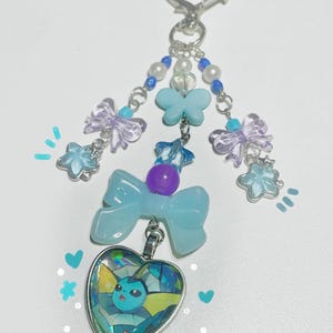May include: A handmade keychain featuring a heart-shaped pendant with a cartoon character design. The keychain includes blue and purple accents, bows, and star-shaped charms. The top has a heart-shaped clasp and beaded accents.