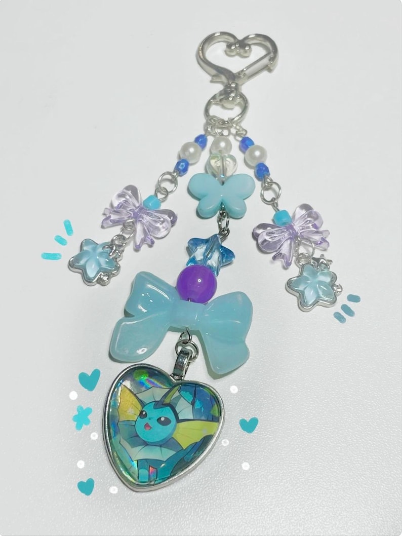 May include: A handmade keychain featuring a heart-shaped pendant with a cartoon character design. The keychain includes blue and purple accents, bows, and star-shaped charms. The top has a heart-shaped clasp and beaded accents.