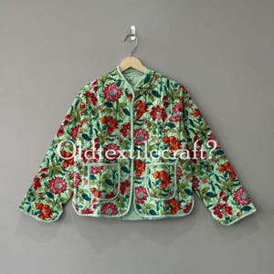 Könnte beinhalten: Eine gesteppte Jacke mit hellgrünem Grund und einem lebendigen Blumenmuster in Rot-, Orange- und Dunkelgrüntönen. Die Jacke hat lange Ärmel, einen Rundhalsausschnitt, zwei aufgesetzte Vordertaschen und eine weiße Einfassung. Der Text Oldtextilecraft? ist sichtbar.