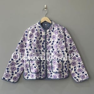 Könnte beinhalten: Eine gesteppte Jacke mit weißem Grund, violettem Blumenmuster und dunkelblauen Rankendetails. Die Jacke hat einen Rundhalsausschnitt, lange Ärmel, eine Knopfleiste auf der Vorderseite und zwei quadratische Taschen mit dunkelblauer Umrandung.