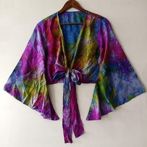 Peut inclure: Un haut court à nouer sur le devant avec de longues manches évasées, orné d'un motif tie-dye aux tons magenta, bleu, vert et jaune. Le vêtement présente un décolleté en V profond et se ferme au centre par deux longs liens en tissu.