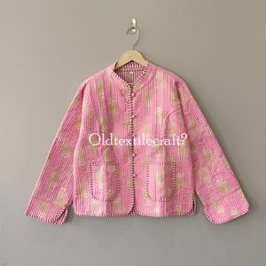 Pode incluir: Um casaco acolchoado em rosa brilhante com um padrão floral dourado repetitivo. O casaco apresenta uma gola mandarim, mangas compridas, dois bolsos frontais e um fecho frontal com botões tipo alamares. As bordas têm costuras visíveis.