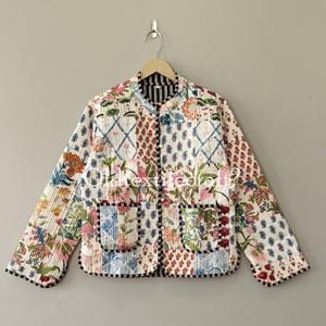 May include: A quilted jacket featuring a white base with a multi-colored floral and geometric block print pattern. The jacket has long sleeves, a round neckline, two front patch pockets, and black and white striped trim edges.