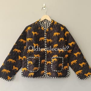 Puede incluir: Una chaqueta acolchada negra con un patrón repetitivo de leopardos amarillos, mangas largas, cuello redondo y dos bolsillos delanteros. Los bordes tienen un ribete a cuadros blancos y negros. El texto Oldtextilecraft? es visible.