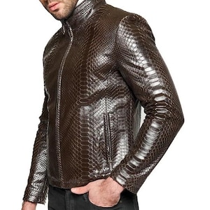 Può includere: Giacca in pelle marrone con motivo a pelle di serpente. La giacca ha una chiusura con cerniera, un colletto rialzato e due tasche anteriori. Le maniche sono lunghe e aderenti. La persona indossa jeans grigio scuro.