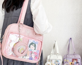 May include: Pink shoulder bag with a clear front pocket displaying colorful anime-style cards and a heart-shaped charm. The bag has a long strap and a small handle. Two other bags, white and lavender, are also visible.