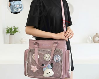 Könnte beinhalten: Eine staubrosa Tragetasche mit einer transparenten Vordertasche, die dekorative Anstecknadeln und einen Plüschcharakter zeigt. Die Tasche hat einen Schultergurt und obere Griffe. Das Bild zeigt auch eine kleinere hellblaue Tasche.