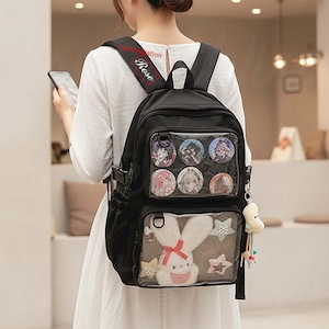 Peut inclure: Sac à dos noir avec plusieurs poches et fenêtres transparentes. Le sac à dos présente un lapin en peluche, des décorations en forme d'étoiles et des épingles de personnages. L'épaule porte le mot "Rose".