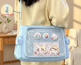 Könnte beinhalten: Eine hellblaue Umhängetasche mit einer transparenten Vordertasche, die dekorative Anstecknadeln und eine Karte mit Cartoon-Illustrationen zeigt. Die Tasche hat einen langen Riemen und Seitentaschen. Das Wort "Rose" ist auf dem Riemen aufgedruckt.