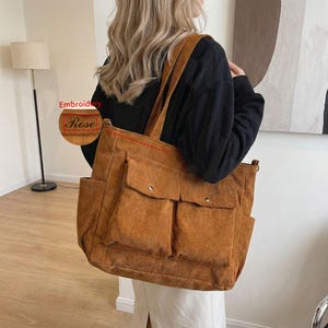 May include: A corduroy tote bag in a warm brown hue, featuring two front pockets and side pockets. The bag has wide shoulder straps and a red embroidered "Rose" detail. The bag is designed for carrying items.