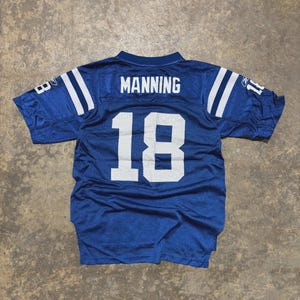 Op de afbeelding: Blauw voetbalshirt met witte strepen op de schouders en het nummer 18 op de rug. De naam "MANNING" staat boven het nummer gedrukt. Het shirt is van mesh-materiaal.