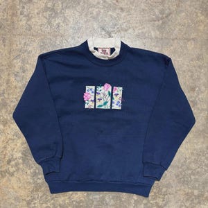 以下が含まれることがあります： 白い襟と花の刺繍が施されたネイビーブルーのスウェットシャツ。 刺繍は、ピンクの花と緑の葉が3つの垂直の長方形のパネルに配置されています。 「Top Stitch」の文字が襟に見えます。