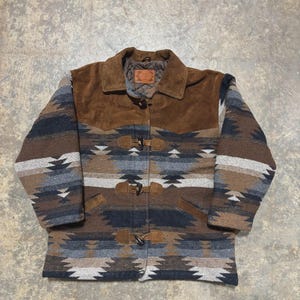 May include: A patterned jacket with a brown suede yoke and collar. The body of the jacket features a geometric design in shades of brown, grey, and blue. It has toggle closures and a quilted lining.