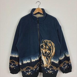 Peut inclure: Veste polaire bleu marine avec fermeture éclair intégrale et col montant. La veste présente un grand motif de loup sur le devant et des motifs décoratifs sur les manches. Accrochée à un cintre en métal.