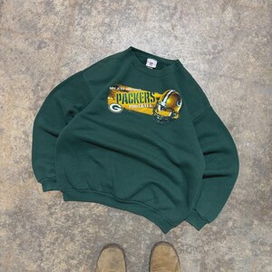 Puede incluir: Sudadera verde de fútbol americano de los Packers con un gráfico de un casco dorado y el nombre del equipo. La sudadera tiene el texto "NOT JUST FOOTBALL" encima del nombre del equipo. Es un estilo de cuello redondo.