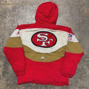 Może przedstawiać: Czerwona, biała i złota kurtka z kapturem z logo San Francisco 49ers. Kurtka ma duże logo drużyny na plecach, z logo NFL na rękawie. Kurtka wykonana jest z wytrzymałego materiału i posiada kaptur.