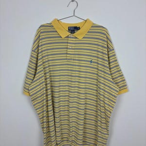 May include: Yellow and navy blue striped polo shirt with a classic collar and short sleeves. The shirt features a small embroidered logo on the chest and is hanging on a metal hanger.
