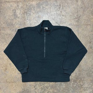 May include: A dark green, long-sleeved sweatshirt with a quarter-zip closure. The sweatshirt has a high collar and ribbed cuffs and hem. The fabric appears to be a thick knit material. The garment is laid flat on a neutral-toned surface.