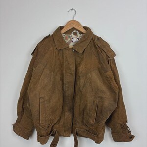Peut inclure: Veste en cuir marron avec col et épaulettes, suspendue à un cintre en bois. La veste présente deux poches avant, une ceinture et une fermeture boutonnée. La doublure a un motif.