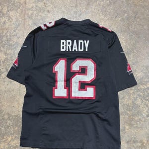 May include: Black football jersey with the name "BRADY" and the number "12" in white with red trim. The jersey has the team logo on the sleeves. The jersey is made of a breathable material.