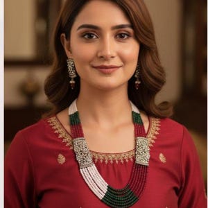 Puede incluir: Una mujer con un vestido rojo y un collar de múltiples hilos. El collar presenta cuentas blancas, verdes y rojas con cierres plateados ornamentados. Pendientes a juego completan el conjunto.