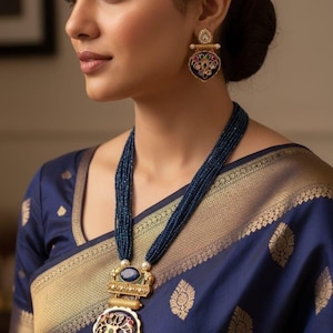May include: A woman wearing a navy blue beaded necklace with a gold pendant and matching earrings. The pendant and earrings feature intricate designs with colorful accents. The woman is also wearing a navy blue sari with gold detailing.