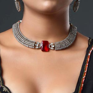 Puede incluir: Un conjunto de collar y pendientes plateados. El collar presenta una gran gema roja rectangular en el centro, rodeada de piedras claras más pequeñas. Los pendientes tienen un diseño similar. Las joyas se lucen sobre un fondo oscuro.