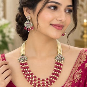 May include: A multi-strand necklace featuring alternating rows of cream-colored pearls and deep pink beads, connected by two ornate, crystal-encrusted floral clasps. Matching drop earrings feature a floral stud with pink and green accents.