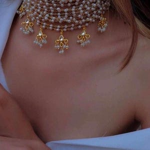 May include: A multi-strand pearl choker necklace with gold-toned accents and dangling pendants. The necklace features small, round pearls and ornate gold-colored charms. The design is elegant and suitable for special occasions.