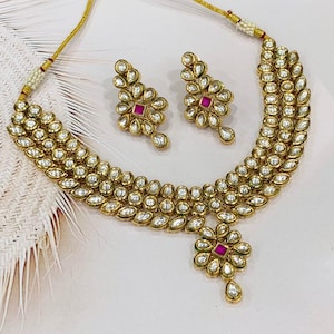 Puede incluir: Un conjunto de collar y pendientes de color dorado. El collar presenta varias filas de pequeñas piedras ovaladas transparentes. Los pendientes y el colgante del collar tienen un diseño floral con una piedra roja central y detalles claros en forma de lágrima. Las joyas se exhiben sobre una superficie blanca.