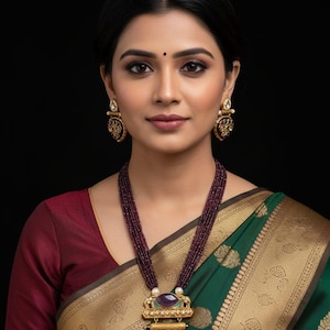 May include: A woman wearing a traditional Indian necklace and earrings. The necklace has a multi-strand beaded chain with a large pendant. The pendant has a gold-coloured frame with a purple stone and red and green enamel detailing. The woman is wearing a green and gold sari.