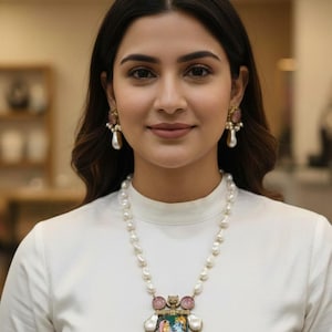 Könnte beinhalten: Eine Frau trägt ein weißes Oberteil, eine Perlenkette und passende Ohrringe. Die Halskette hat einen Anhänger mit einem farbenfrohen Bild und rosa Edelsteinen. Die Ohrringe haben ein ähnliches Design mit Perlen und rosa Akzenten.