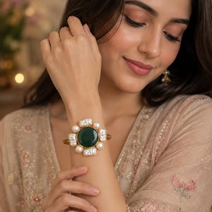 May include: A gold-toned cuff bracelet featuring a large, circular, dark green central stone surrounded by a cluster of irregular, white, oblong-shaped pearls and smaller round white pearls arranged in a symmetrical floral-inspired pattern.