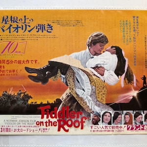 May include: A vintage movie poster for "Fiddler on the Roof" features a man carrying a woman, with text in English and Japanese. The poster has a warm color palette with a scene of horsemen in the background.