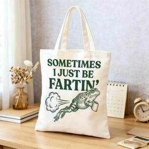 May include: A cream-coloured canvas tote bag featuring dark green text that reads "SOMETIMES I JUST BE FARTIN'" above a detailed illustration of a frog holding a roll of toilet paper with a cloud graphic emanating from behind the frog.