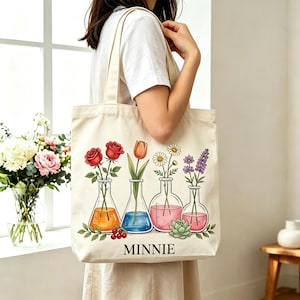 May include: A cream-colored canvas tote bag featuring a graphic print of four glass laboratory flasks containing flowers. The flasks hold red roses, an orange tulip, a white daisy, and purple lavender. The name MINNIE is printed in black below.