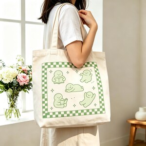 May include: A cream-coloured tote bag with a green chequered border and a design featuring four cartoon figures in various poses. The figures are light green and surrounded by small star shapes. The bag is held by a person.