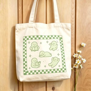 May include: Cream tote bag with a green and white chequered border. The bag features a green illustration of cartoon figures in various poses, including reading and relaxing. The bag has two cream-coloured handles.