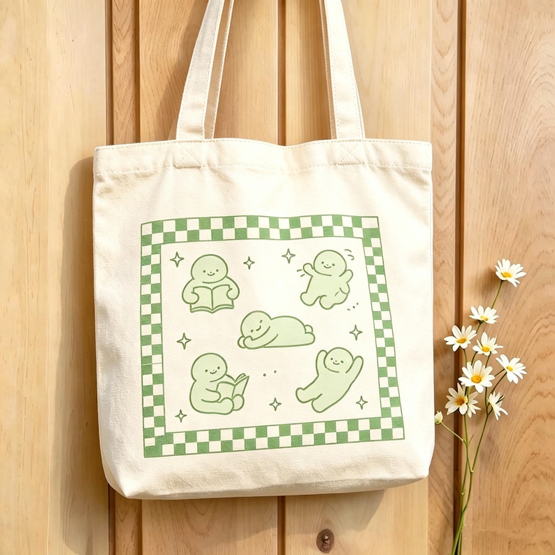 May include: Cream tote bag with a green and white chequered border. The bag features a green illustration of cartoon figures in various poses, including reading and relaxing. The bag has two cream-coloured handles.