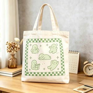 May include: Cream canvas tote bag featuring a green and white checkered border with five green cartoon figures. The bag has two beige handles. The tote bag is on a wooden surface.