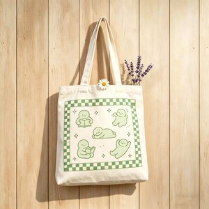 May include: Off-white tote bag featuring a green chequered border and cartoon figures. The bag has long handles and is decorated with a daisy and lavender sprigs. The figures are in various poses, including reading and relaxing. The bag is hanging on a wooden wall.