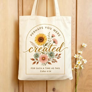 Könnte beinhalten: Cremefarbene Tragetasche mit floralem Design und dem goldenen Schriftzug "Perhaps You Were Created For Such A Time As This". Das florale Design umfasst eine Sonnenblume, rosa und grüne Blumen und braune Blätter.