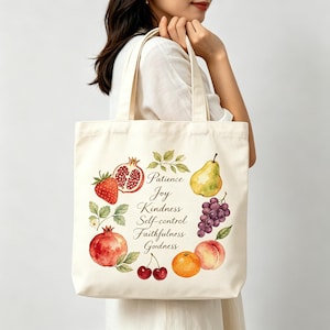 Puede incluir: Bolso tote de color crema con ilustraciones de acuarela de varias frutas, incluyendo fresa, granada, pera, uvas, cerezas, naranja y melocotón. La bolsa también tiene las palabras "Paciencia, Alegría, Bondad, Autocontrol, Fidelidad, Bondad."